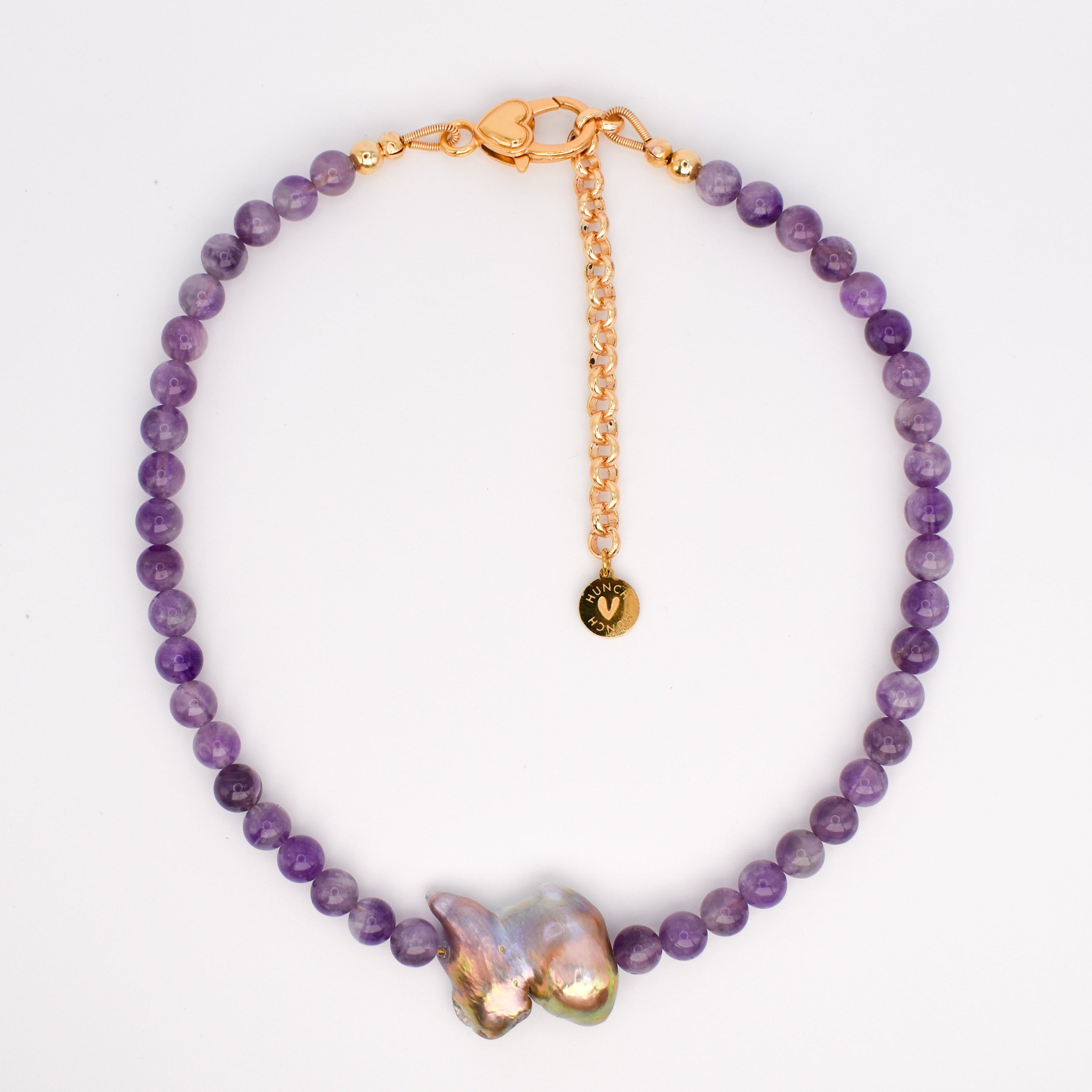 A beaded bracelet featuring purple amethyst beads, a gold-tone chain extender with a small round charm, and two baroque shell accents in a gold-tone setting, displayed on a white background.