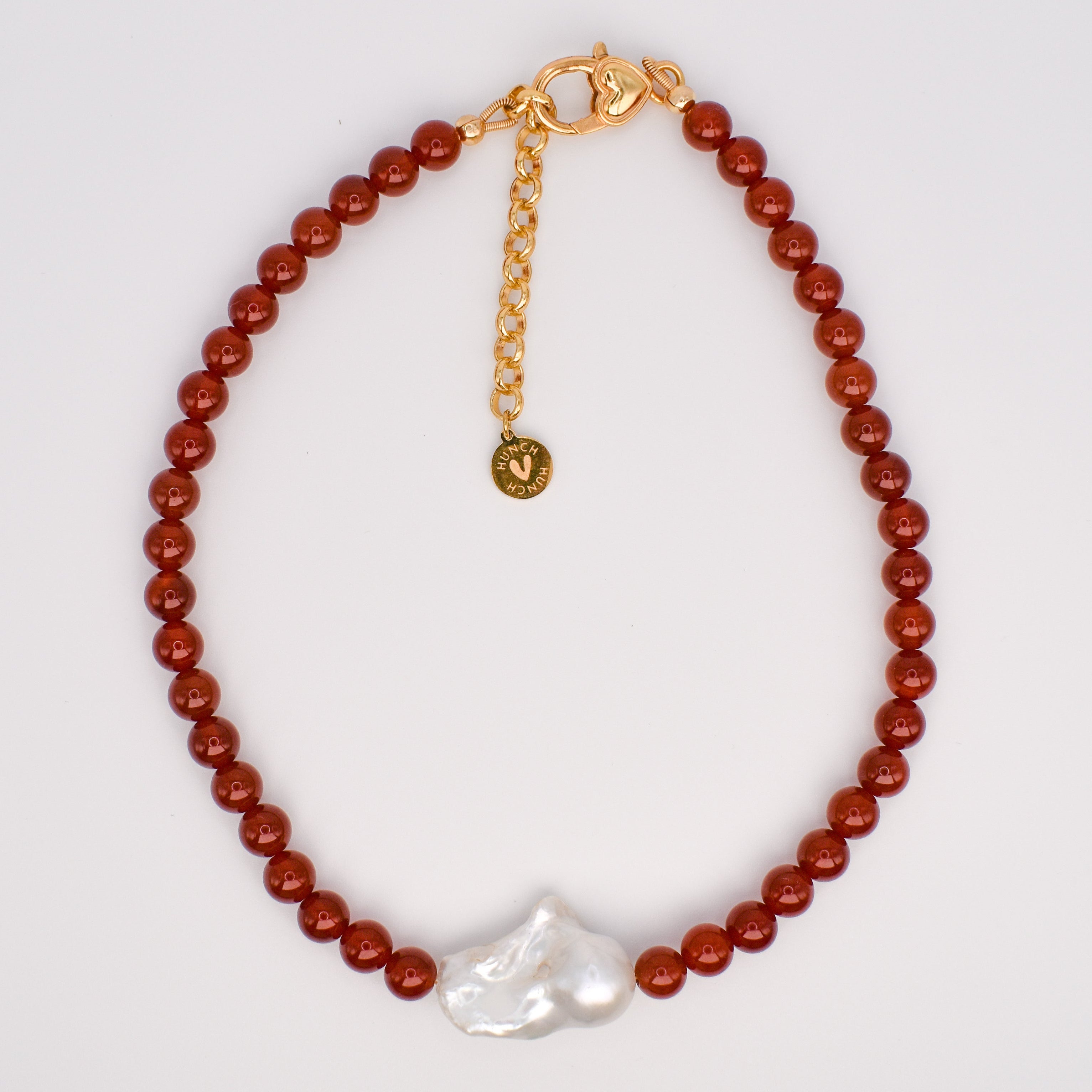 A beaded bracelet featuring round reddish-brown beads, a gold-tone chain extender with a small round charm, and a single irregular white baroque pearl at the bottom, displayed on a white jewelry stand.