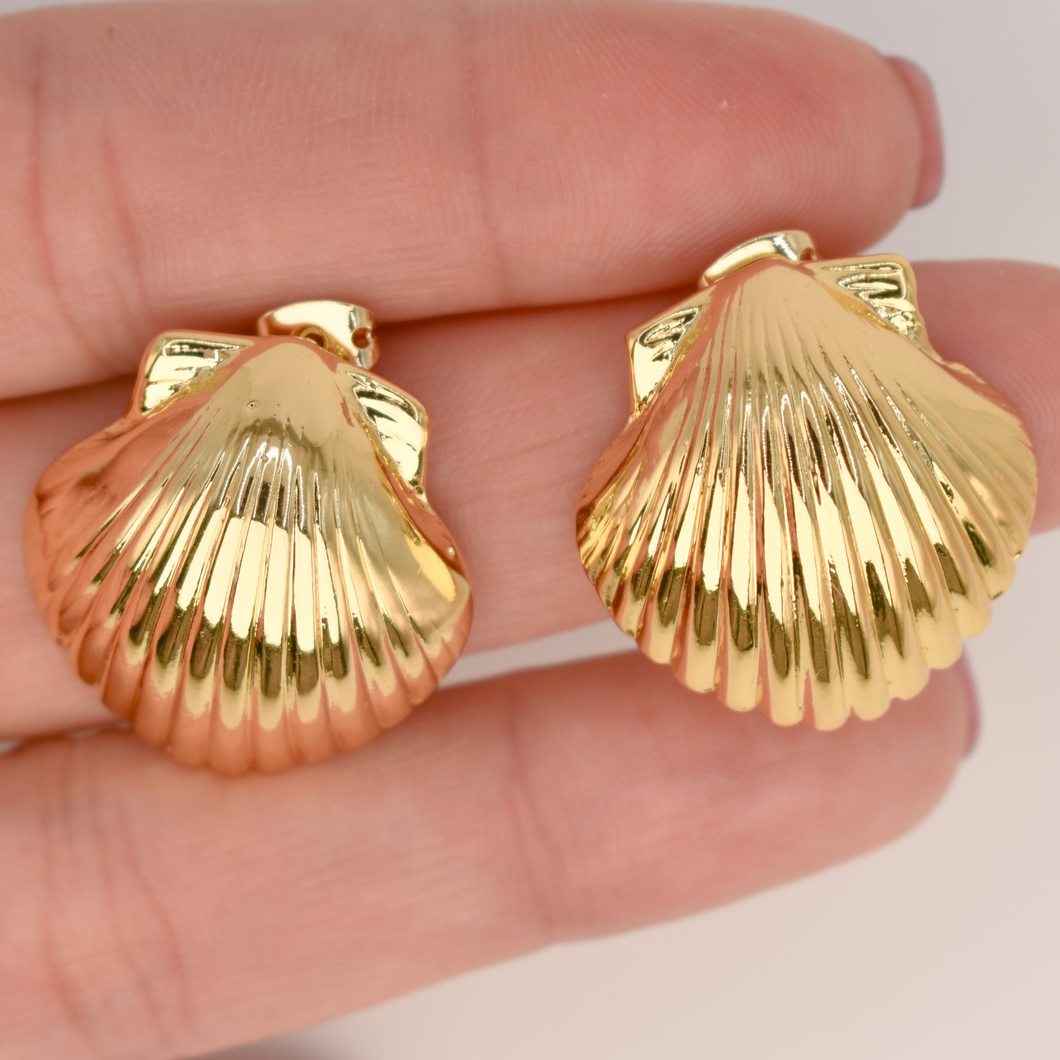 A pair of gold-toned shell-shaped earrings held on a person's fingers against a plain light background.